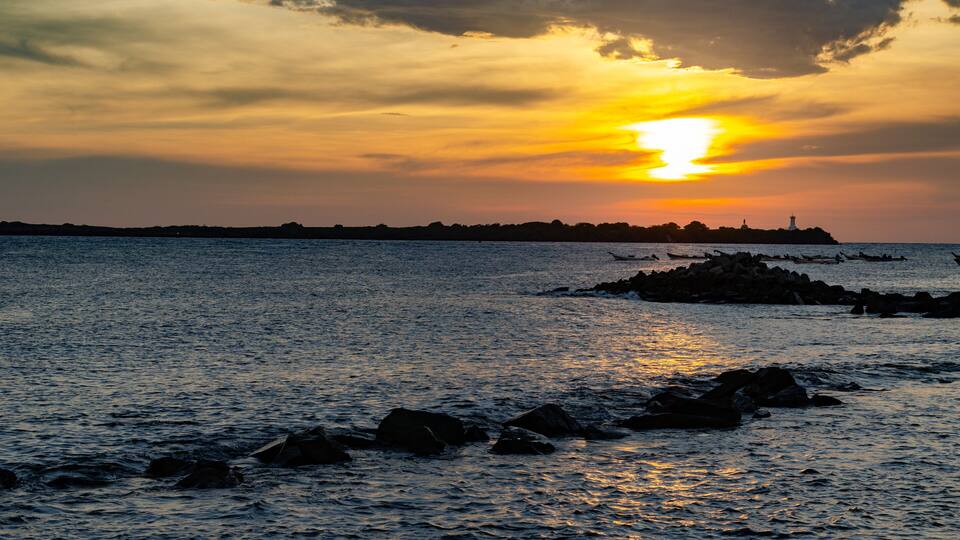 A sunset over a calm sea with rocks in the foreground and a distant shoreline with a lighthouse.