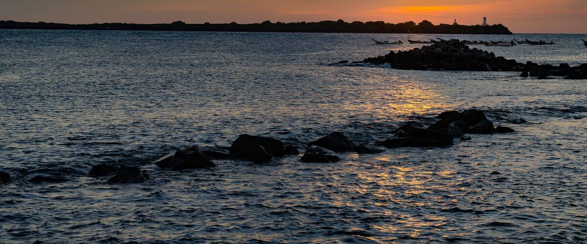 A sunset over a calm sea with rocks in the foreground and a distant shoreline with a lighthouse.