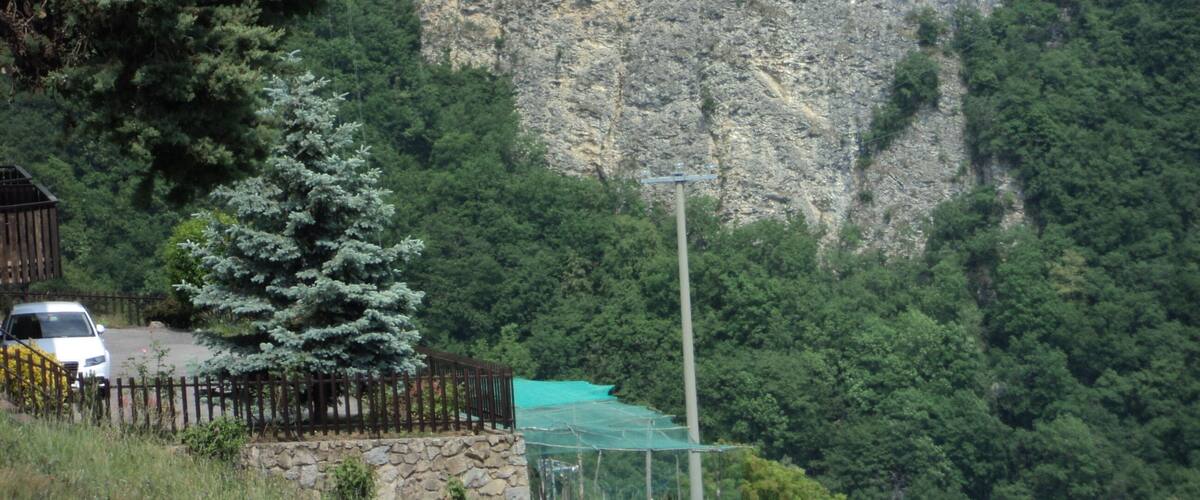 A mountainous landscape with a rocky cliff, dense forest, and a fenced area with a white car.