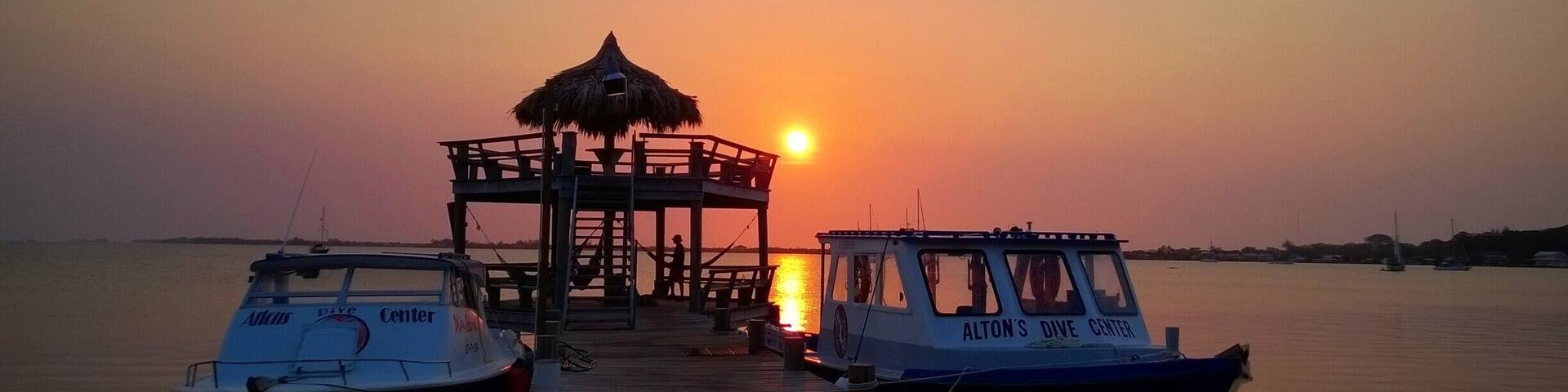 A sunset over a calm body of water with a pier leading to a thatched-roof structure. Two boats are docked at the pier, one labeled 'Alton's Dive Center' and the other 'NAIDA J'.