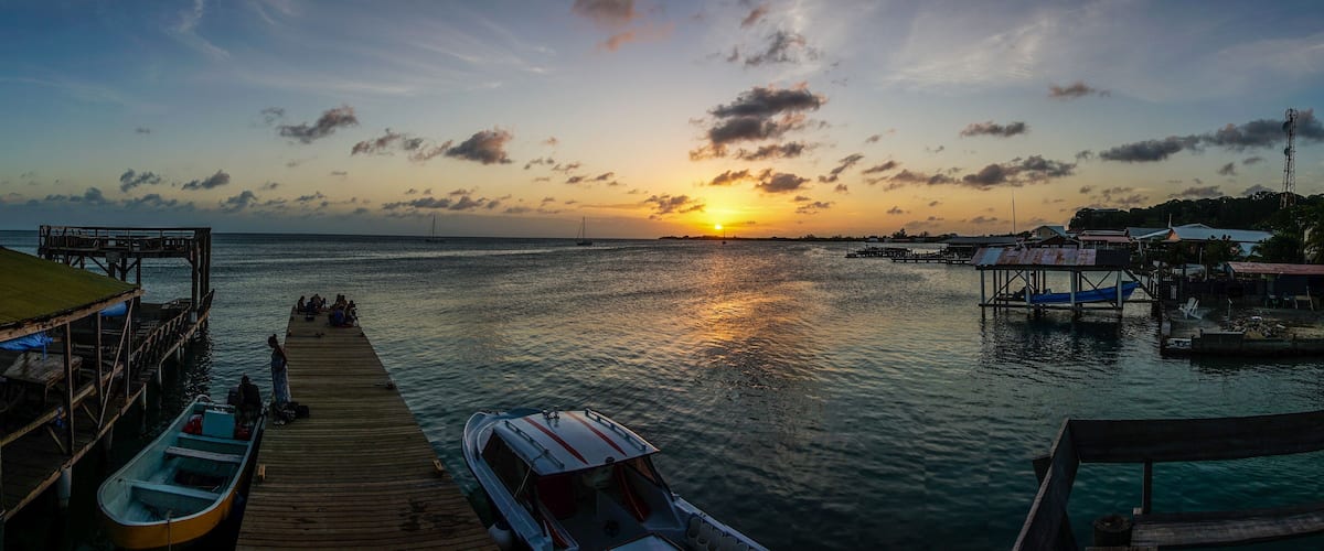 A sunset over a calm sea with a pier, boats, and a distant sailboat.