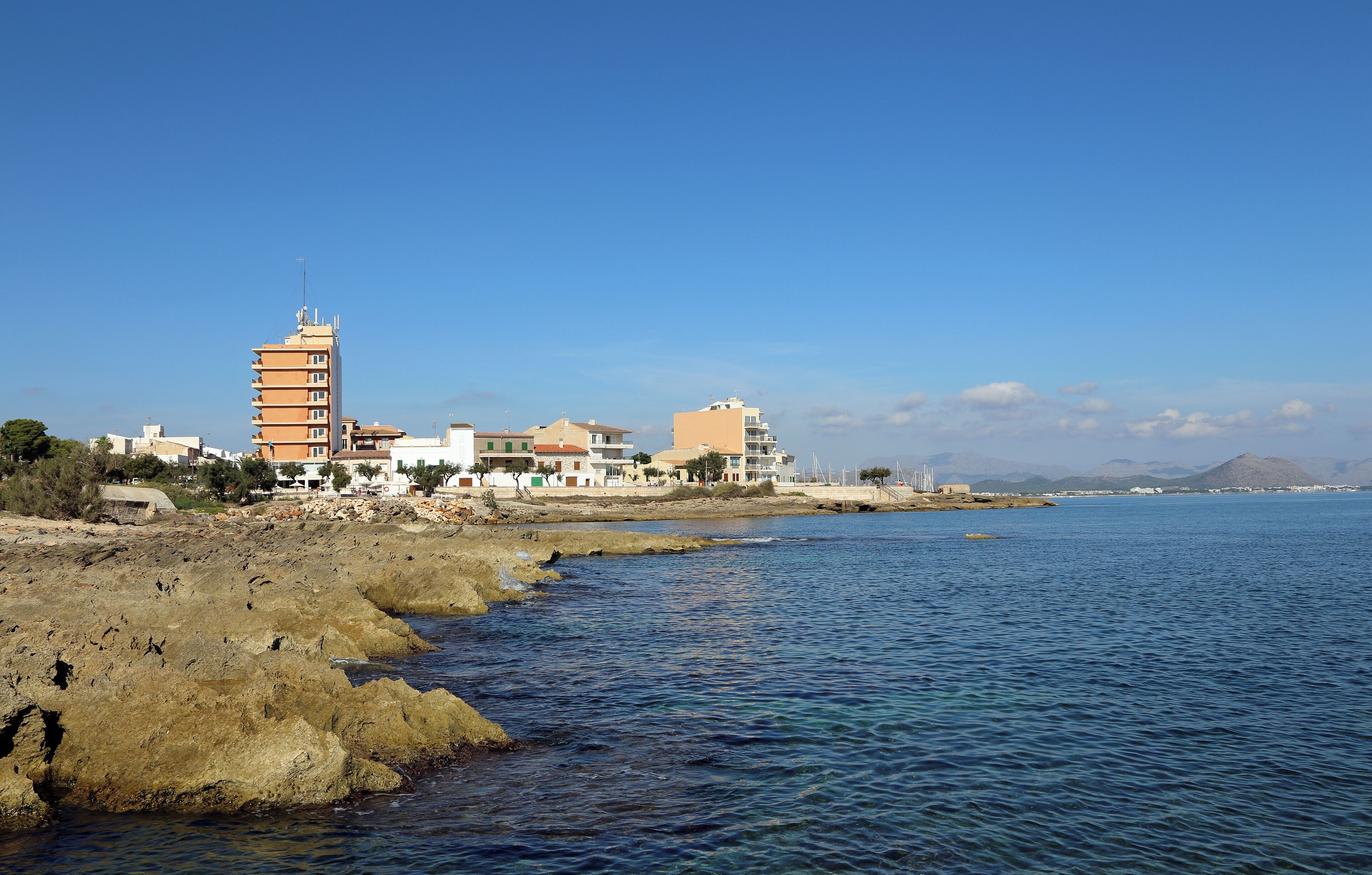 A coastal town with buildings along the shore, rocky terrain, and a clear blue sky.