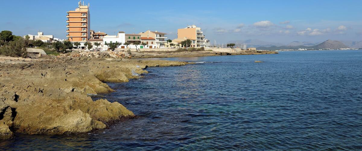 A coastal town with buildings along the shore, rocky terrain, and a clear blue sky.