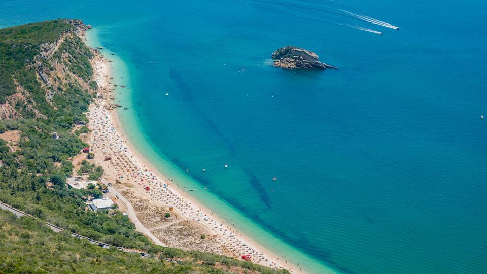 An aerial view of a coastal area with a beach, clear turquoise waters, a small island, and a few boats.