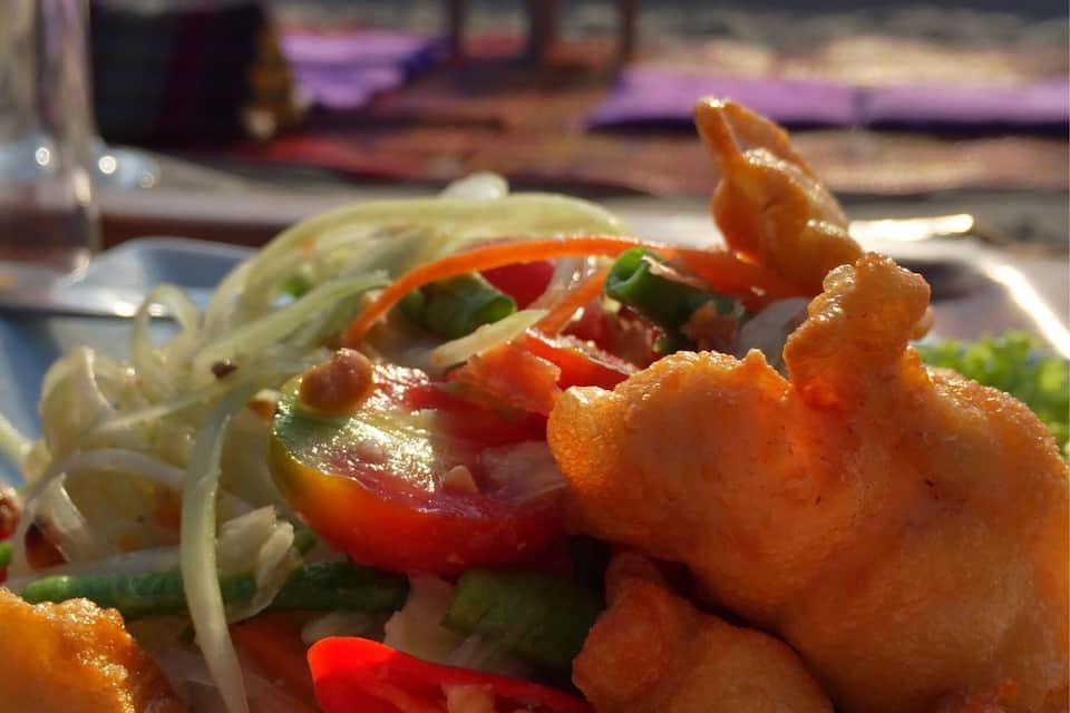A plate of food with noodles, vegetables, and fried items, with a beach and ocean in the background.