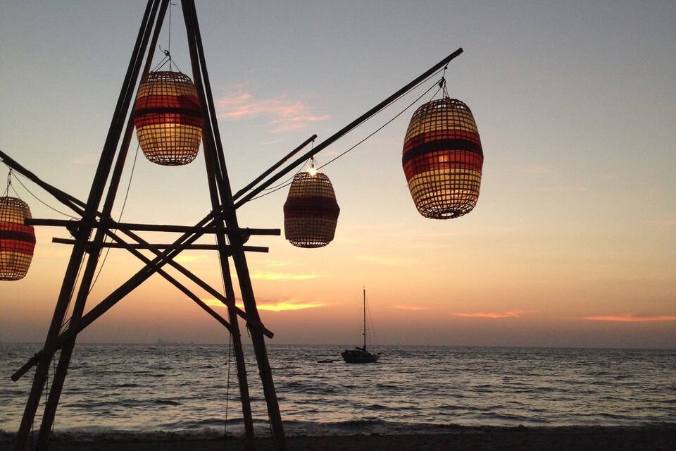 A beach at sunset with a sailboat in the distance and decorative hanging lanterns.