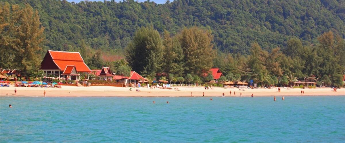 A beach with red-roofed buildings and a forested hill in the background.
