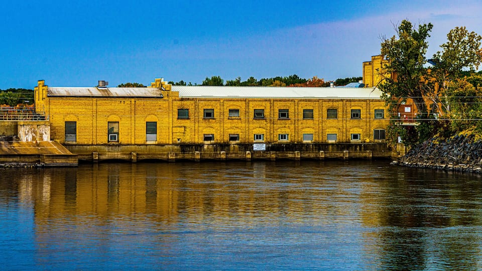 A yellow brick building with a white roof by a calm river.