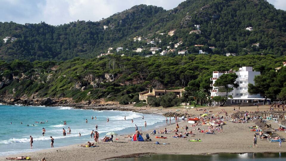 A crowded beach with people sunbathing and swimming, surrounded by lush green hills and buildings.
