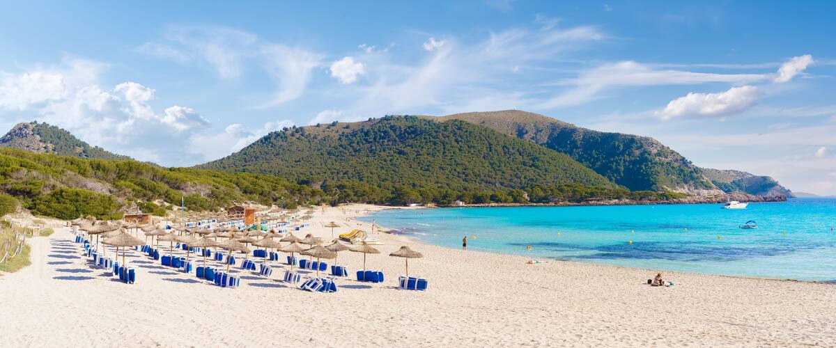 A beach with umbrellas, a clear blue sea, and a mountain in the background.