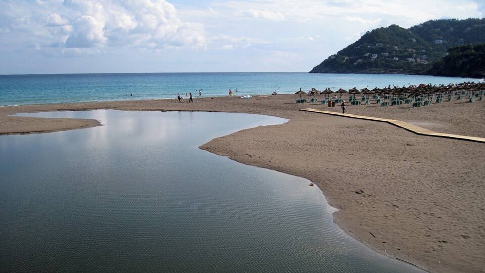 A beach with calm waters, a sandy shore, and a hilly landscape in the background.