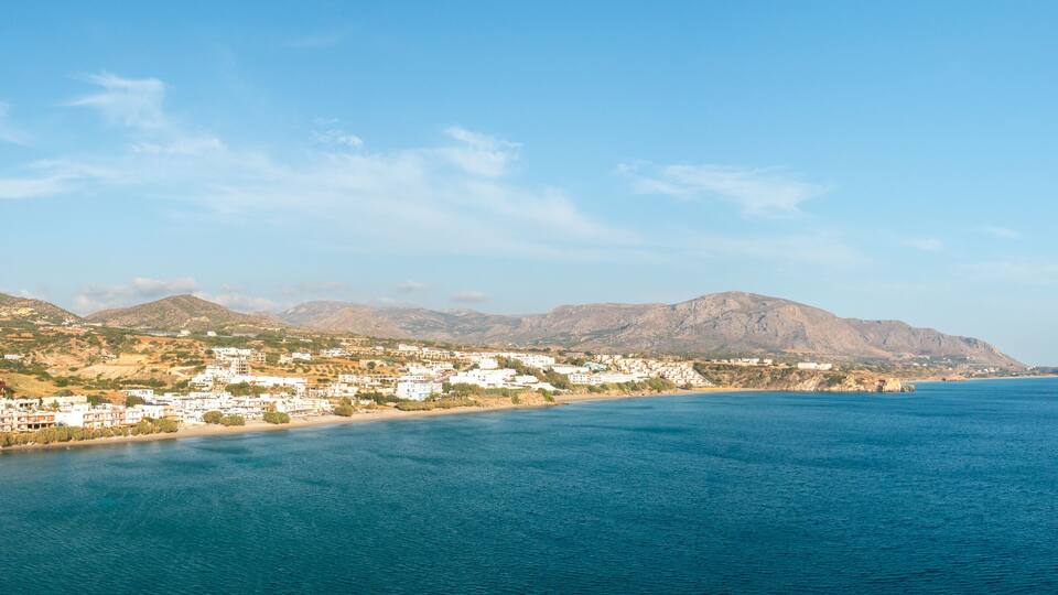 A coastal town with white buildings, a sandy beach, and mountains in the background.