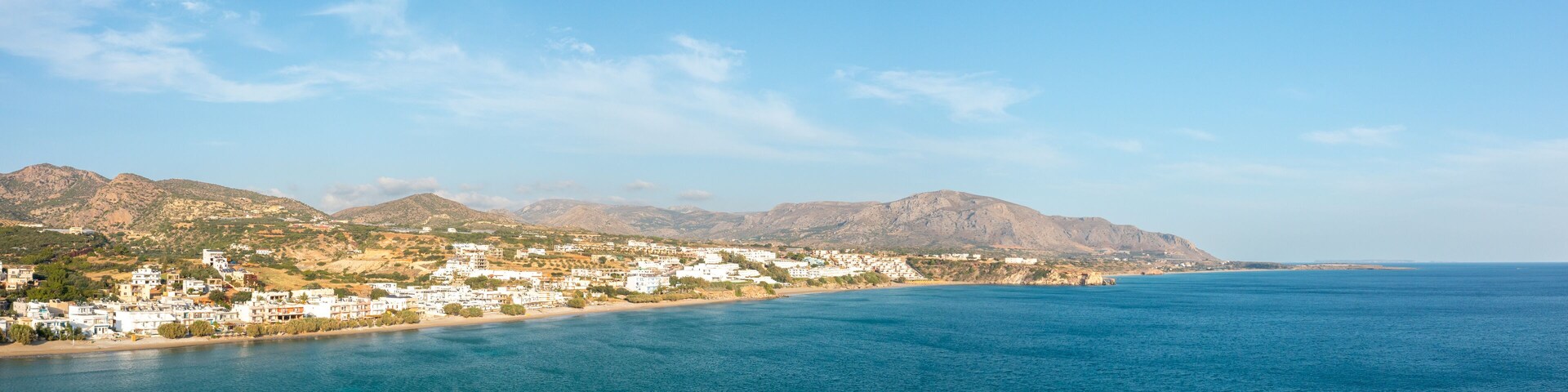 A coastal town with white buildings, a sandy beach, and mountains in the background.