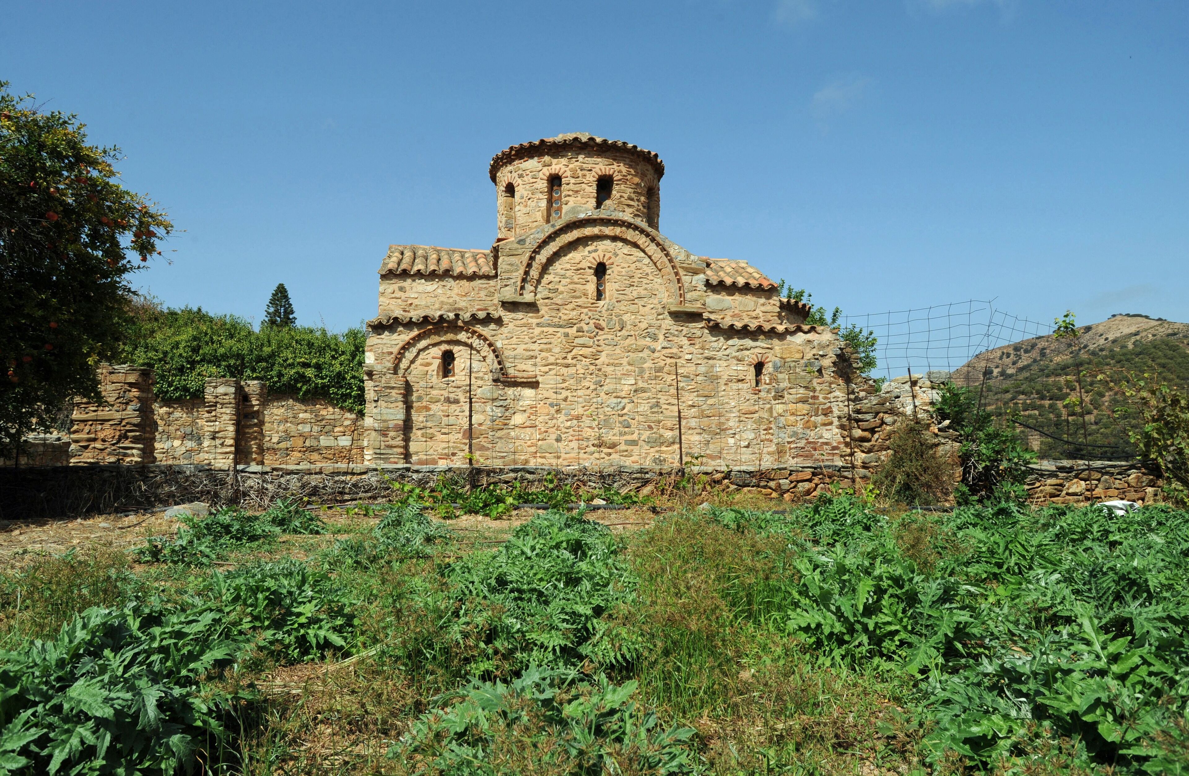 An ancient stone church with a bell tower, surrounded by greenery and hills.