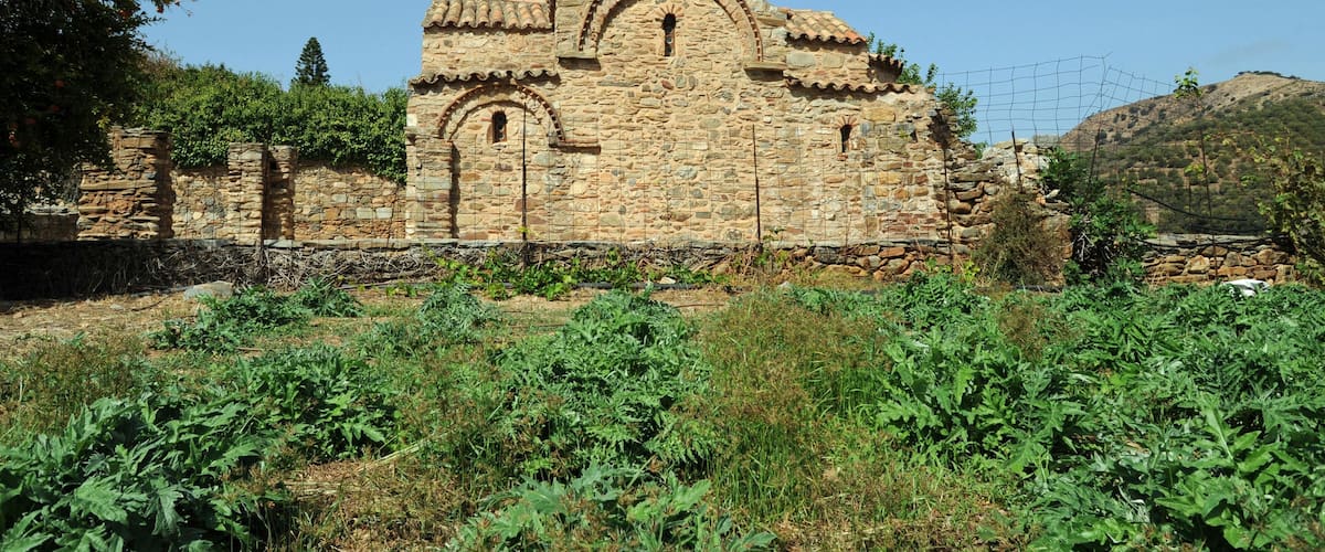 An ancient stone church with a bell tower, surrounded by greenery and hills.