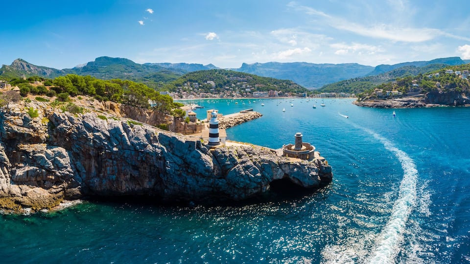 A rocky island with a lighthouse, surrounded by clear blue waters and a mountainous coastline.