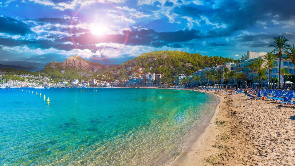 A beach with clear turquoise water, a sandy shore, and a backdrop of buildings and a mountain.