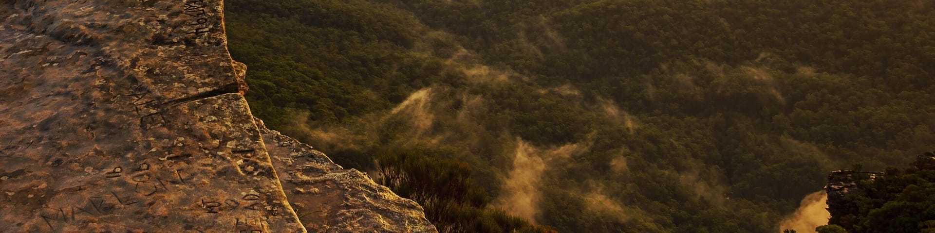 A person standing on a cliff overlooking a valley with mountains in the distance during sunset.