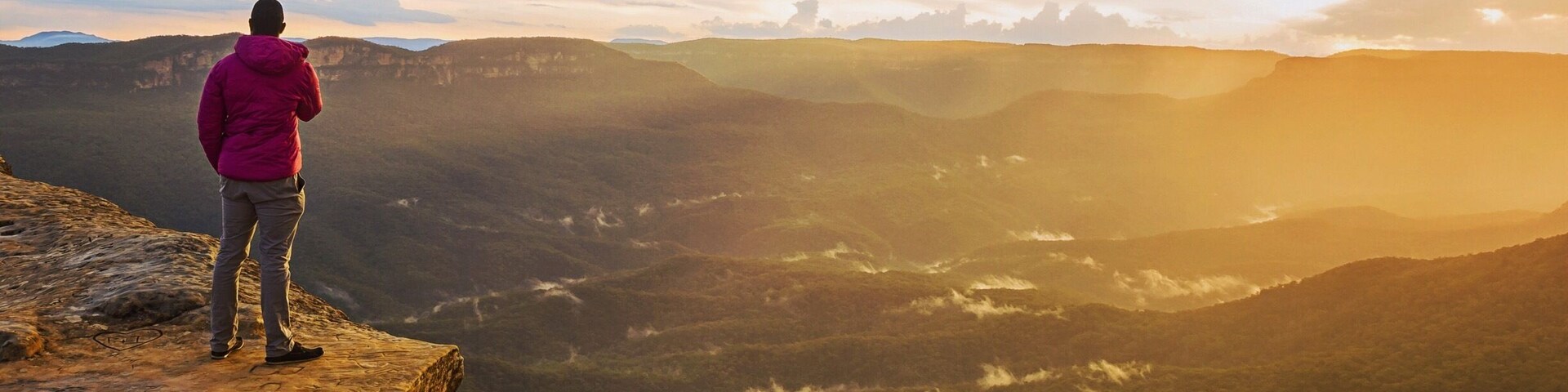 Una persona parada en un acantilado, con vistas a un valle y montañas distantes al atardecer.