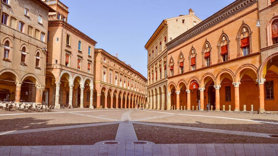 A historic square with a central open space, lined with buildings featuring arched walkways and red awnings.