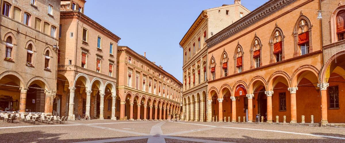 A historic square with a central open space, lined with buildings featuring arched walkways and red awnings.