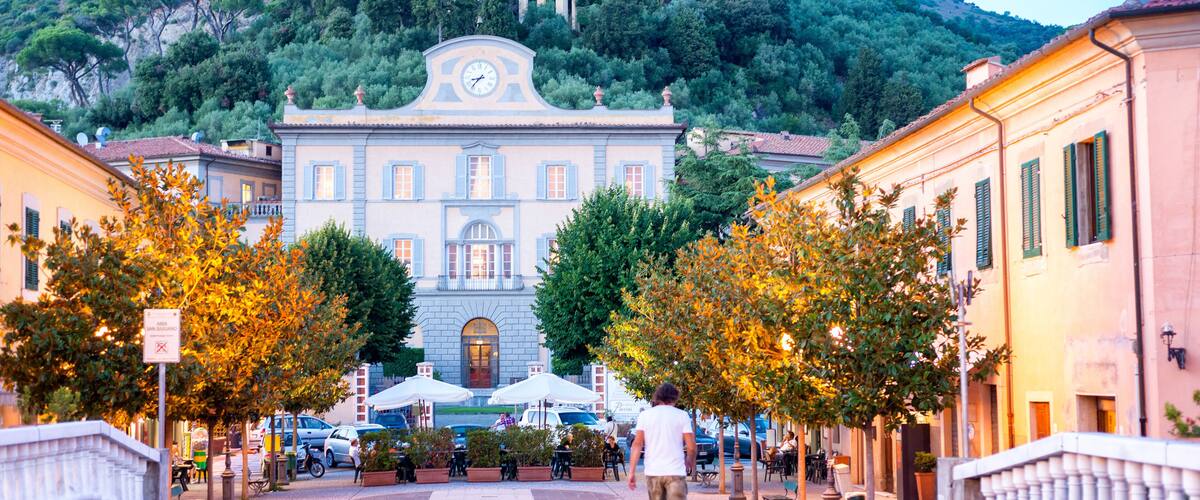 A town square with a prominent building featuring a clock tower, surrounded by trees and cafes.