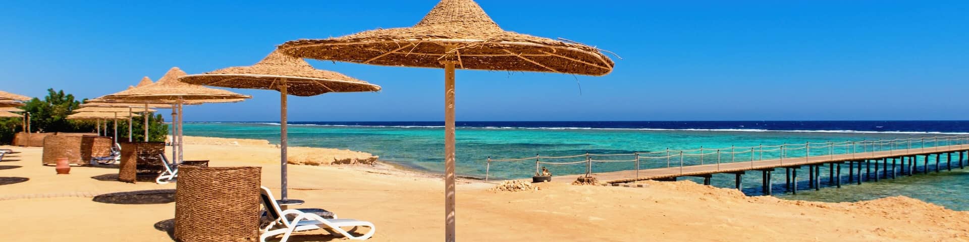 A beach with thatched umbrellas, lounge chairs, and a wooden pier extending into the sea.