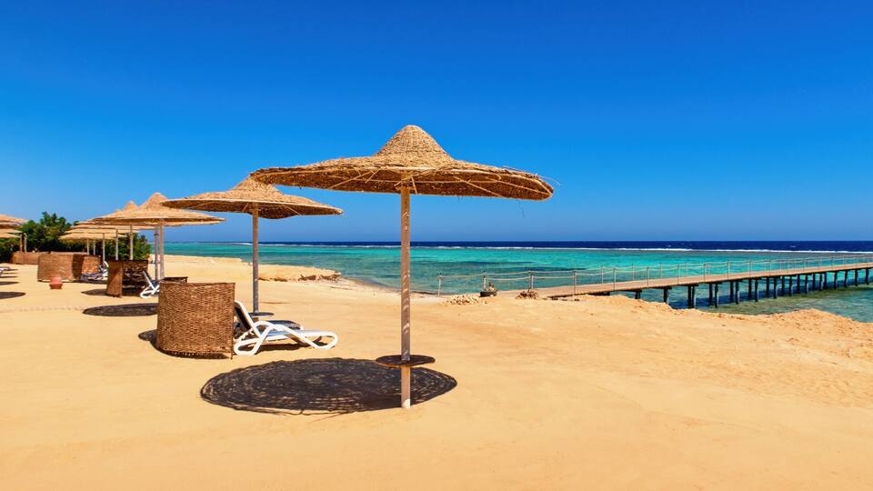 A beach with thatched umbrellas, lounge chairs, and a wooden pier extending into the sea.