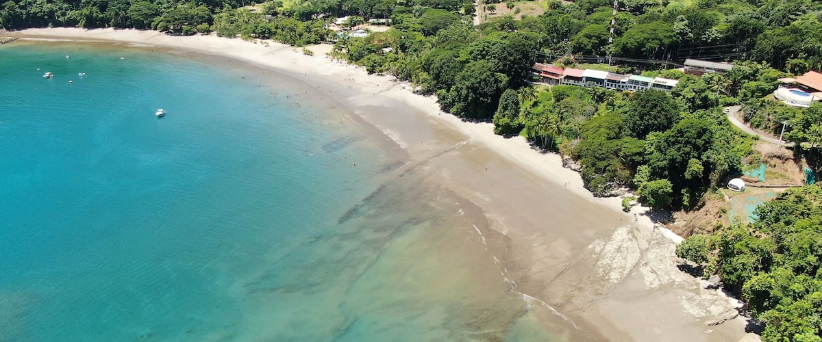 A coastal beach with clear turquoise waters, a sandy shore, and dense green vegetation.