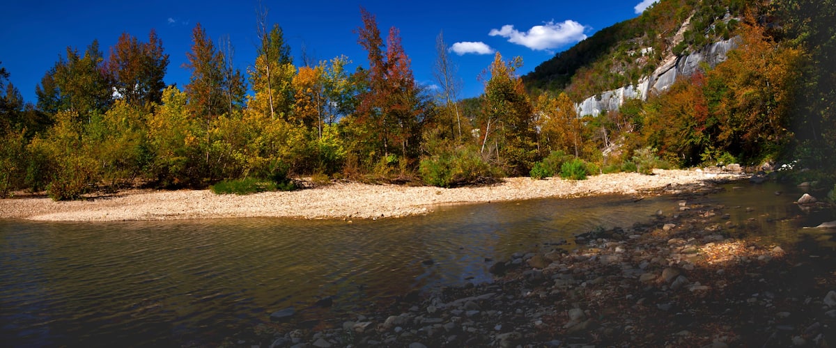A river with clear water, surrounded by autumn-colored trees and a rocky shoreline.