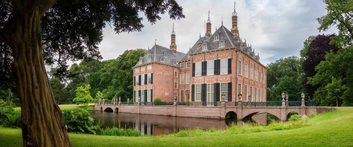 A historic brick building with a large tree in the foreground, a bridge over a canal, and lush greenery in the background.