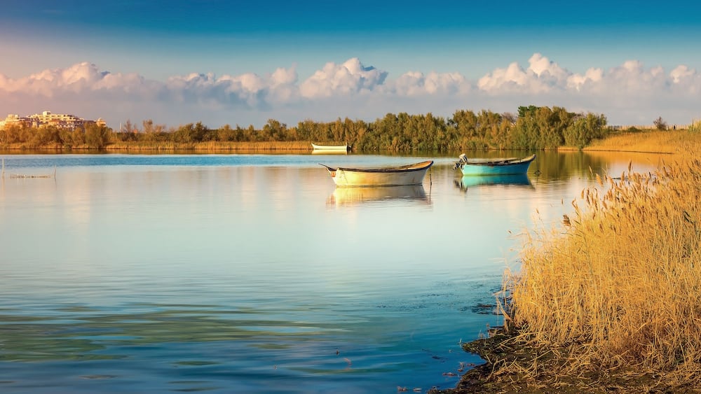 Un lac paisible avec deux bateaux, un bâtiment situé au loin et une rive bordée d’une forêt.