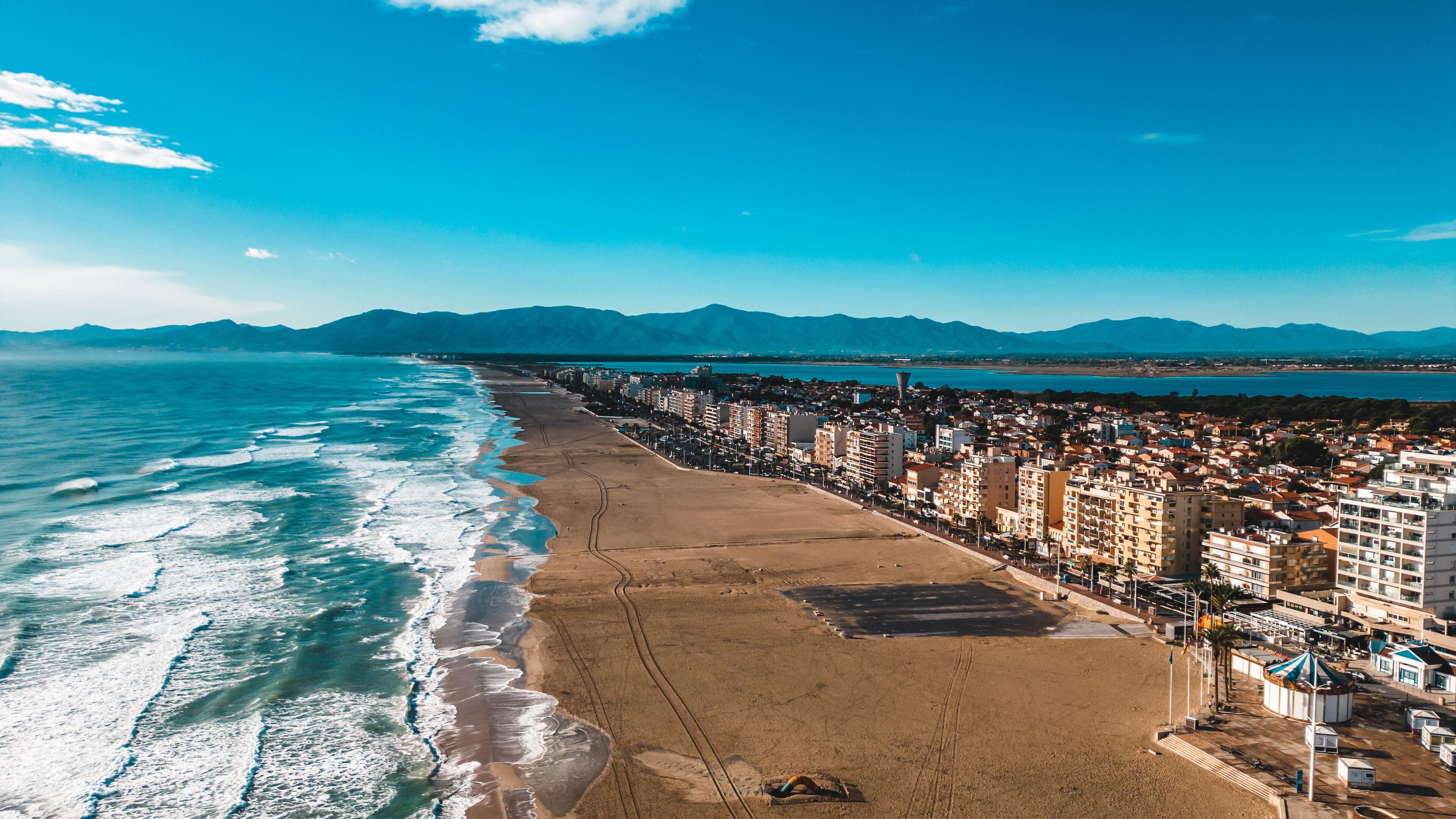 Aerial view of the beach meeting the urban landscape under a bright blue sky, a symphony of sand, sea, and city, Canet-en-Roussillon, Occitanie, France.