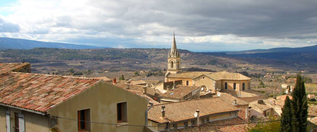 A town with a prominent church tower, terracotta-roofed buildings, and a mountainous backdrop.