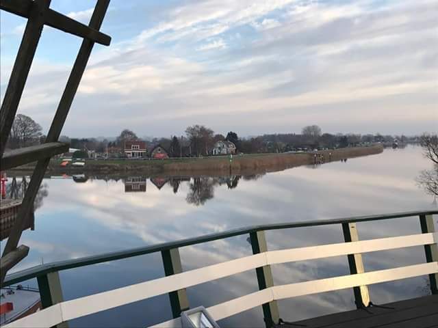 A calm river with a wooden bridge in the foreground and houses along the riverbank in the background.