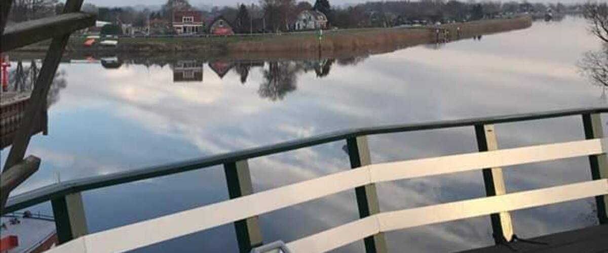 A calm river with a wooden bridge in the foreground and houses along the riverbank in the background.