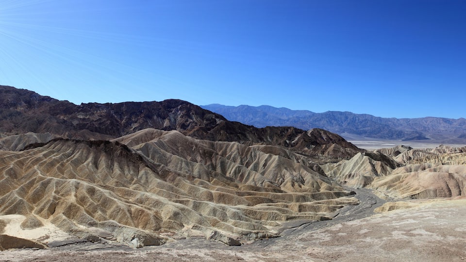 A desert landscape with rugged, eroded hills and distant mountains under a clear blue sky.