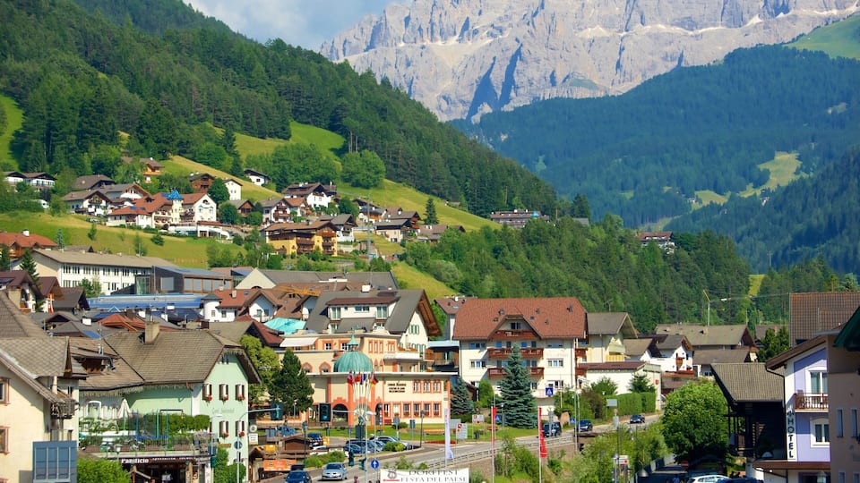 A mountain village with houses on a hillside, a road with cars, and a mountain range in the background.
