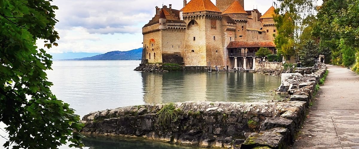 A historic castle by a calm lake, surrounded by lush greenery and a stone pathway.