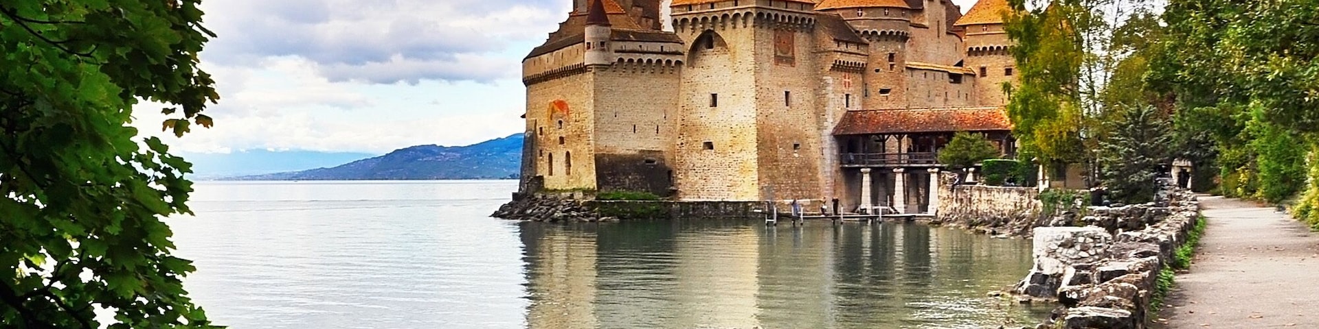A historic castle by a calm lake, surrounded by lush greenery and a stone pathway.