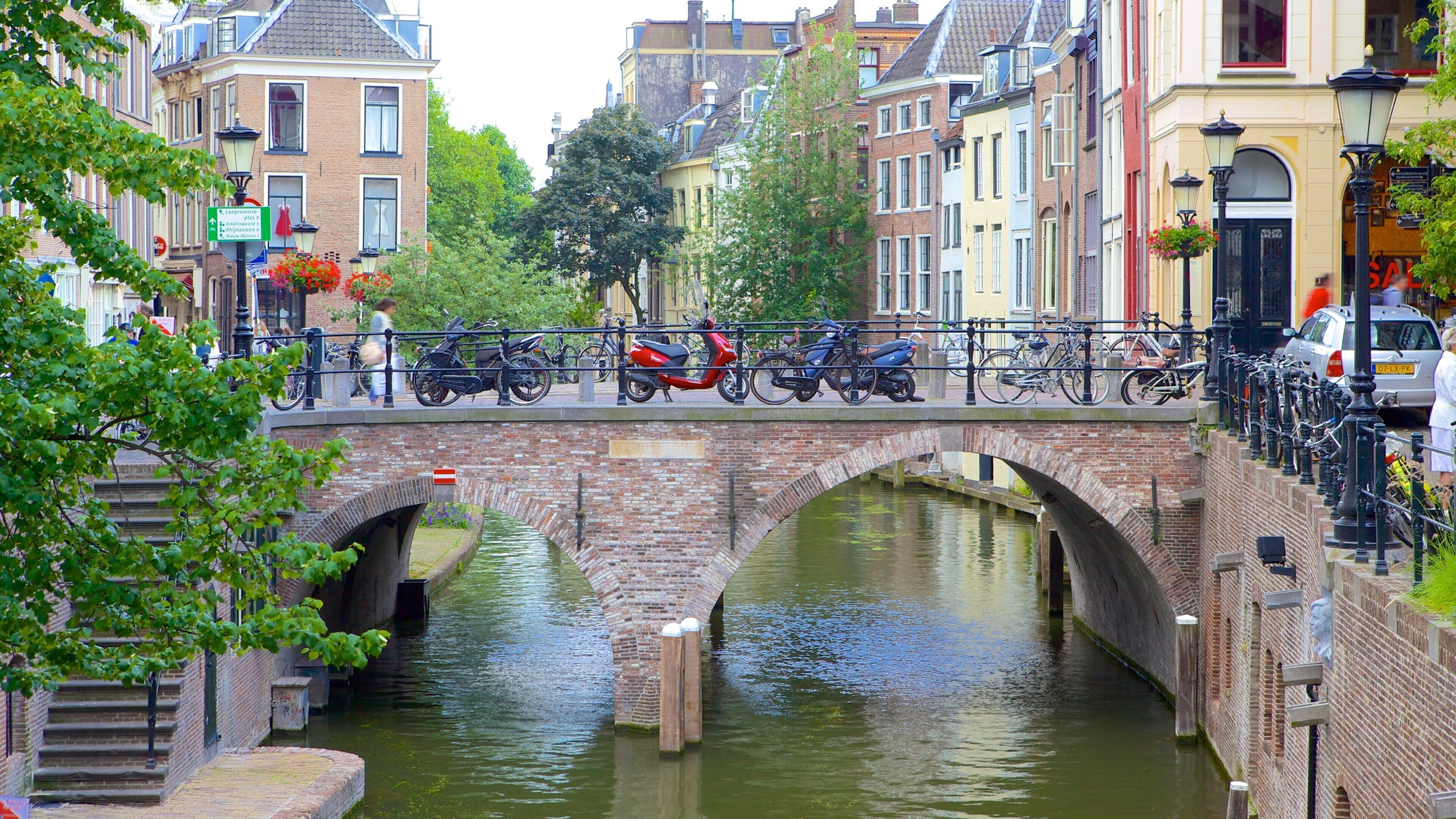Un canal con un puente de piedra, flanqueado por bicicletas y patinetes, y edificios históricos.