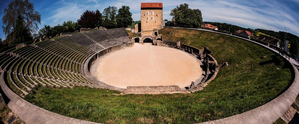 A historic amphitheater with tiered seating and a central stage, surrounded by a grassy area and a small tower.