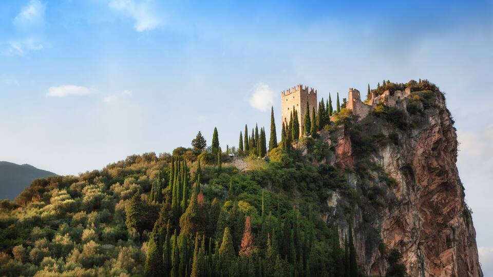 A castle perched atop a rocky cliff, surrounded by dense greenery and cypress trees.