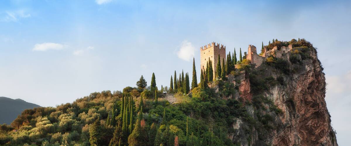 A castle perched atop a rocky cliff, surrounded by dense greenery and cypress trees.