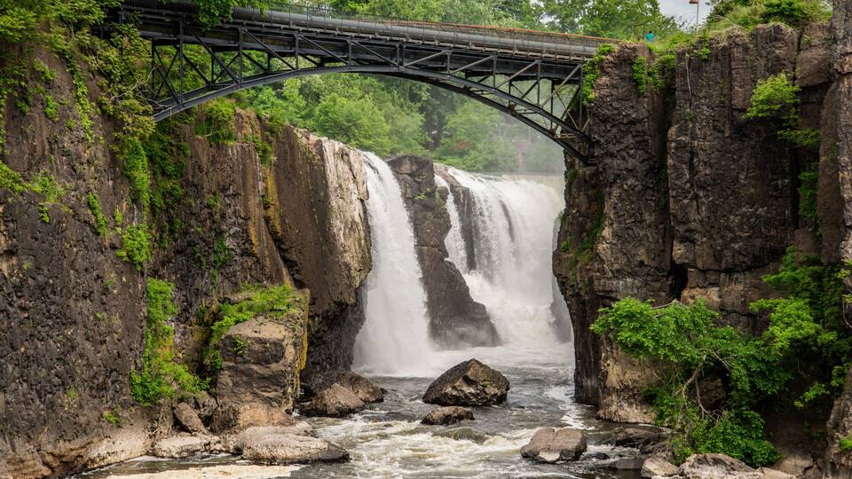 A waterfall under a bridge with rocky cliffs and greenery.
