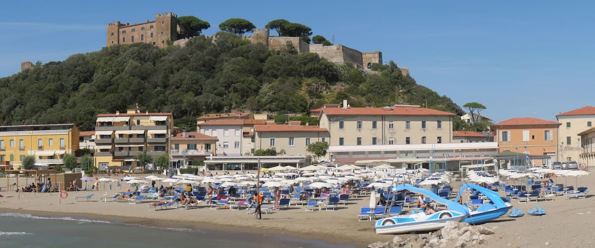 A beach with sun loungers and umbrellas, a castle on a hill, and buildings in the background.