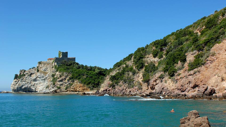 A coastal landscape with a castle on a cliff, clear blue sea, and rocky terrain.