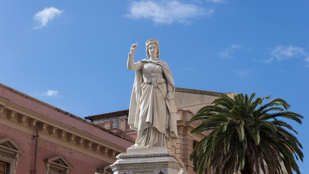 A statue of a woman with a raised hand, a palm tree, and a building with arched windows.