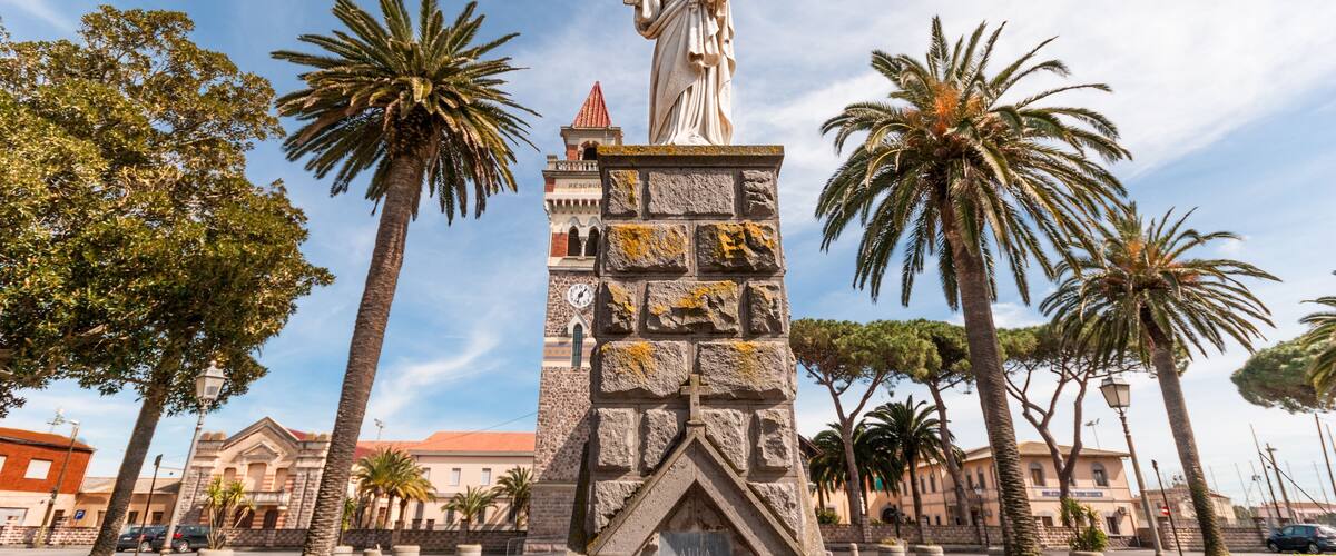 Un monumento con una estatua, palmeras y un edificio con una torre del reloj al fondo.