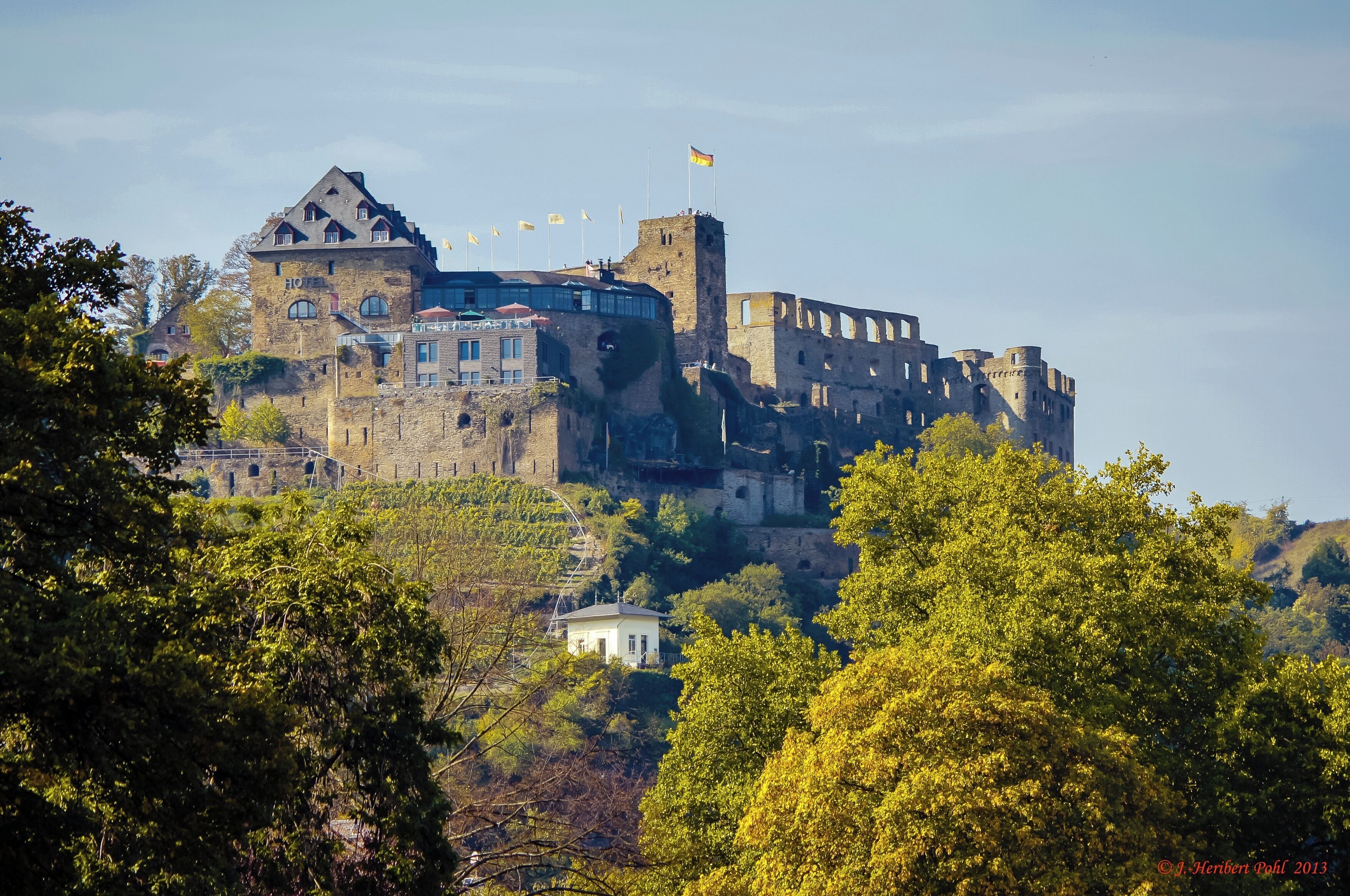A historic castle with multiple towers and flags, surrounded by lush greenery and trees.